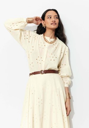 Femme portant un chemisier crème à œillets et une jupe avec ceinture marron, collier chaîne en or, grandes boucles d'oreilles en forme de fleurs, et larges bracelets dorés, posant devant un mur blanc.