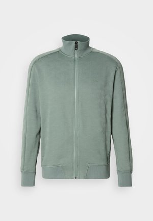 Veste zippée verte avec un col montant, présentant un motif géométrique texturé et un détail de logo. Fabriquée en tissu doux, avec des poignets côtelés.