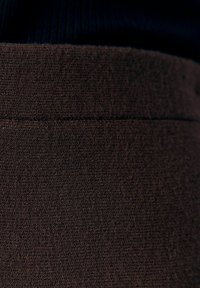 Tissu brun avec une surface texturée, probablement tissé, présentant une couture sur le bord supérieur, entourée de tissu sombre en haut.