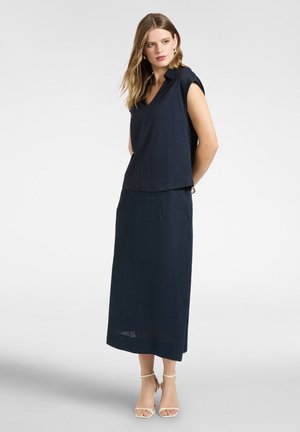 Kvinde med lyst brunt hår iført en ærmeløs navy top og matchende langt nederdel, kombineret med hvide åbne tå hæle, stående med hænderne bag ryggen.