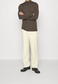 Pull en col montant marron, pantalon large crème et chaussures noires, mettant en avant un mélange de tissus doux et une coupe ajustée.