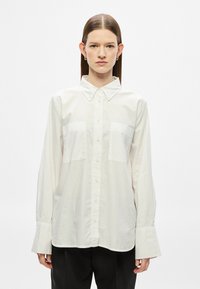 Fehér hosszú ujjú gombos ing hegyes gallérral, két mellzsebbel és széles mandzsettával. Simább, könnyű anyagból készült.