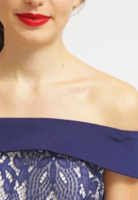 Marinblå off-shoulder klänning med en strukturerad halsringning, med spetsdetaljer och blommönster samt texturerad underlag.