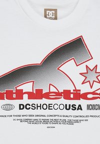 Bílá košile s velkým logem DC Shoes v černé a červené, text "DCSHOECOUSA" a motivační značková zpráva dole.
