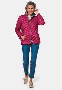 Fuchsia gewatteerde jas met hoge kraag en ritssluiting, voorzien van een chevronpatroon en zijzakken, gecombineerd met blauwe jeans en beige sneakers.