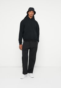 Črna kapuca s kenguru žepom, v kombinaciji s črnimi trenirkami z belimi stranskimi črtami. Nosite jo s črno vedro kapo in belimi supergami.