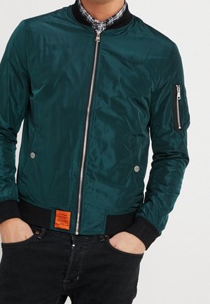 Mann trägt eine türkisfarbene Bomberjacke mit schwarzen Bündchen und Kragen, orangefarbenem Etikett nahe der Taille, Hände in den Taschen, über einem karierten Hemd.