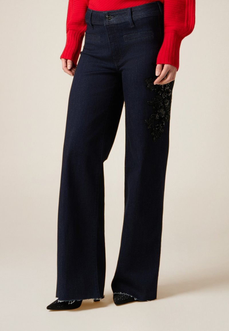 Donkerblauwe wijde jeans met een gestructureerde zwarte bloemige kraalversiering op de linker dij en rafelige zomen, gecombineerd met een rood topje.