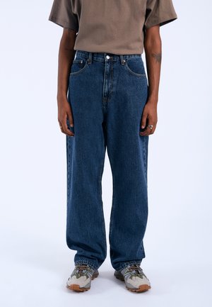 Dark blue denim jeans with a loose fit, featuring front pockets, stitched seams, and a button closure. Worn with gray and green sneakers.