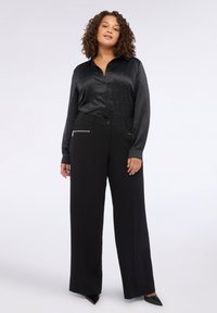 Camisa de satén negra de botones con pequeñas chispas, combinada con pantalones anchos negros que tienen bolsillos con cremallera y una textura suave.