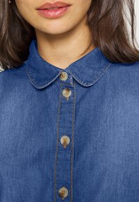 Denimskjorta i ljusblå färg, klassisk krage och knäppning med beige sömmar och runda, pärlliknande knappar.