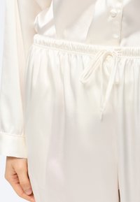 Κοντινό πλάνο ενός ατόμου που φοράει παντελόνι από ελεφαντόδοντο σατέν με ελαστική μέση και δεμένο κορδόνι, συνδυασμένο με ένα ταιριαστό σατέν πουκάμισο με κουμπιά.