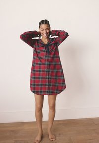 Červené flanelové šaty s károvaným vzorom a zelenými detailmi, zapínané na gombíky vpredu, s dlhými rukávmi a zaobleným spodným lemom. Pri golieri je tmavá kravata.