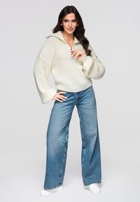 Pull en tricot crème avec manches cloche et col à demi-fermeture éclair, associé à un jean en denim bleu à jambes larges et des baskets blanches.