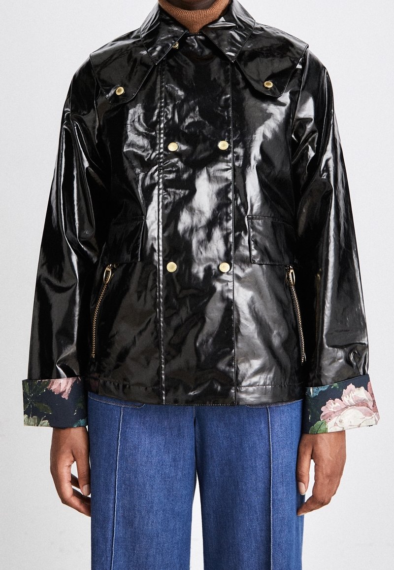 Veste noire brillante à double boutonnage avec boutons dorés, doublure à fleurs aux poignets, portée sur un col roulé marron et un jean bleu.