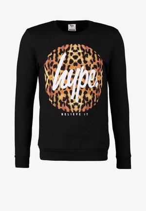 Hombre con camisa negra de manga larga con estampado de leopardo y texto, vaqueros gris claro, botas marrones, llevando una bolsa de deporte beige con asas marrones.