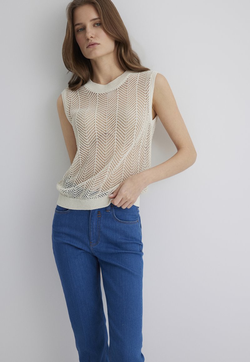 Ärmelloses Strickoberteil in Creme mit einem Zickzackmuster, kombiniert mit blauen Jeans aus Denim, die fünf Taschen und einen Knopfverschluss haben.