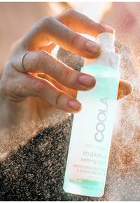 Klarer, hellgrüner Flakon von COOLA Make-up-Setting-Spray mit einem Sprühkopf, der in einer Hand gehalten wird und von einem feinen Nebel umgeben ist. 1,5 Fl. Oz.