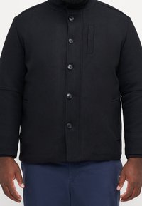 Czarna kurtka z mieszanki wełny z wysokim kołnierzem, zapięciem na guziki z przodu, bocznymi kieszeniami oraz dyskretną kieszenią na piersi. Gładka faktura, strukturalny krój.