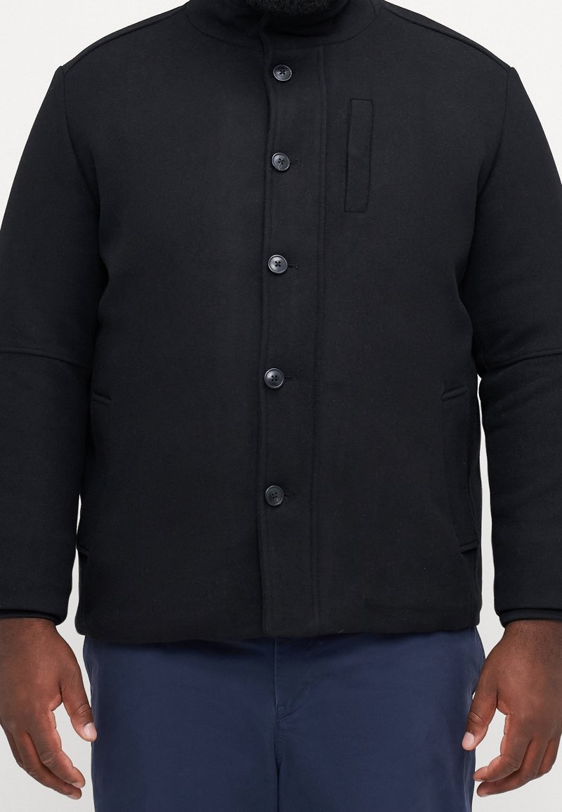 Czarna kurtka z mieszanki wełny z wysokim kołnierzem, zapięciem na guziki z przodu, bocznymi kieszeniami oraz dyskretną kieszenią na piersi. Gładka faktura, strukturalny krój.
