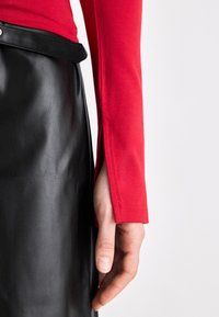 Rotes Langarmoberteil mit eng anliegender Textur, kombiniert mit einem hoch taillierten schwarzen Lederrock mit seitlichem Schlitz und glatter Oberfläche.