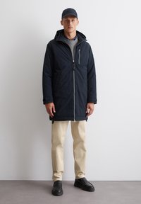 Námornícky modrý zateplený parka s kapucňou, predným zipsom a kontrastnými detailmi na zips. Obsahuje dlhé rukávy a bočné vrecká. Nosené nad béžovými nohavicami.