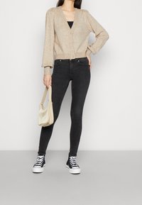 Cárdigan de punto beige con mangas abullonadas, cuello en V y delantero con botones, combinado con jeans skinny negros y zapatillas altas azul marino, sosteniendo un bolso crema.