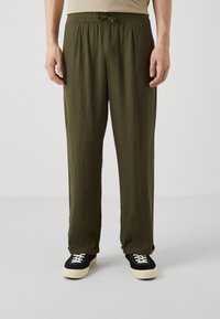 Olijfgroene, lichte broeken met een elastische tailleband en een trekkoord. Heeft een relaxed fit en opgerolde manchetten, gecombineerd met zwarte sneakers.