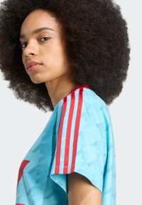 Camisa desportiva azul com um padrão texturizado, detalhes em riscas vermelhas nas mangas e um grande logótipo gráfico à frente.