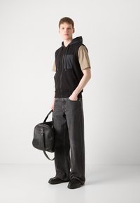 Μαύρο γιλέκο με φερμουάρ και γκρίζα τσέπη, μπεζ κοντομάνικο πουκάμισο, σκούρο γκρι φαρδύ τζιν και μαύρη τσάντα με λουράκι. Περιλαμβάνονται μαύρα παπούτσια.