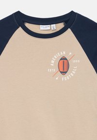 Camisa de mangas raglan bege e azul-marinho com gola redonda e gráfico laranja de futebol com o texto "American Football ESTD 1999" no peito.