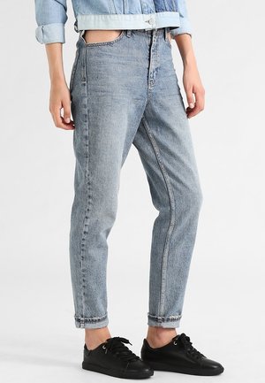 Persoon draagt lichtblauwe opgerolde jeans en zwarte sneakers, staand tegen een effen witte achtergrond.