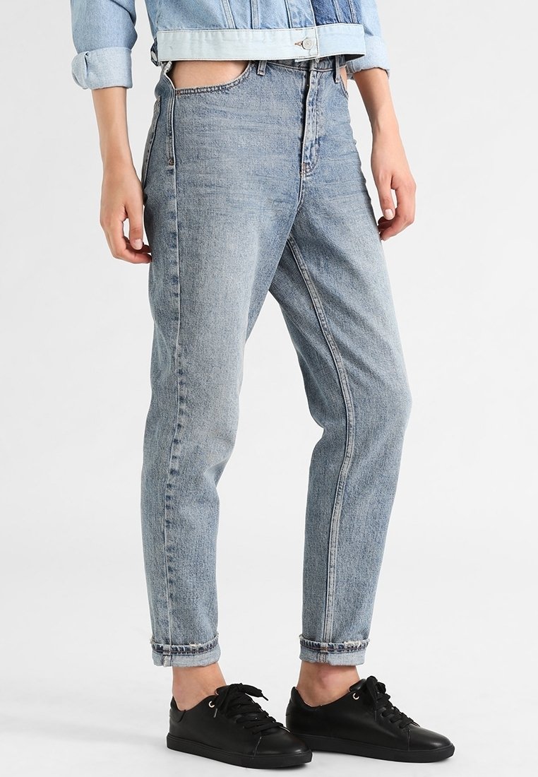 Personne portant un jean bleu clair retroussé et des baskets noires, debout devant un fond blanc uni.