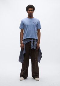 Ανοιχτό γαλάζιο κοντομάνικο μπλουζάκι με λογότυπο κειμένου, καφέ παντελόνι με φαρδιά πόδια και μπλε καρό πουκάμισο δεμένο στη μέση. Ουδέτερο φόντο.