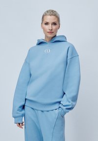 Hanorac de culoare albastru deschis, fabricat dintr-un material moale, având un buzunar tip cangur, manșete ribbate și un logo brodat pe piept.