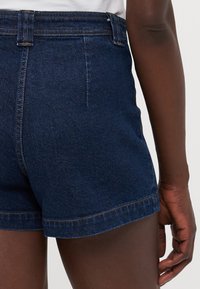 Mørke denimshorts med høy midje, med rullekant, synlig søm og en jevn tekstur. Bakside som viser lomme detaljer.