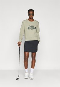 Φωτεινό ελαιώδης πράσινος φούτερ με γραφικό κάστρου, συνδυασμένος με μια μαύρη φούστα. Το μοντέλο φορά τυπωμένα γυαλιά ηλίου και λευκά αθλητικά παπούτσια, κρατώντας μια γκολφομπαλά.