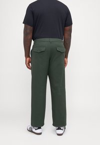 Grüne weite Hose mit Gesäßtaschen, aus weichem Stoff. Getragen mit einem schwarzen T-Shirt und weißen Sneakers mit schwarzen Streifen.