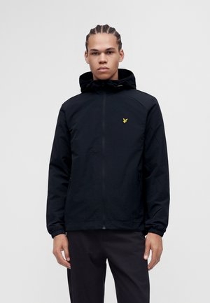 Jeune homme aux cheveux tressés portant une veste à capuche noire avec un petit logo d'oiseau jaune sur la poitrine, debout devant un fond uni.