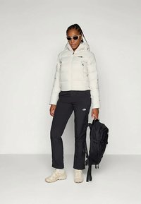 Bela podložena jakna s kapuco, črne hlače, bež superge in črn nahrbtnik. Obleko dopolnjujejo sončna očala.