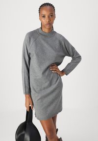 Vestido de malha cinza com mangas longas e um decote alto, apresentando um corte relaxado e fendas laterais. Modelo segura uma bolsa preta.