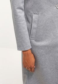 Manteau sweat-shirt gris présentant une texture de tissu lisse, des manches longues, et une poche latérale avec un détail de couture discrète.