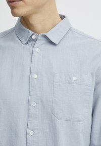 Lys blå skjorte med knapper laget av myk stoff med krage, med hvite knapper og lomme på venstre bryst.