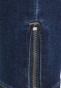 Jeans de denim azul escuro com um fecho lateral em metal prateado e costuras amarelas ao longo da costura.