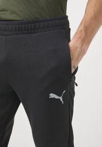 Melni sporta bikses ar gludu tekstūru, ar aukliņu vidukļa joslu un sudraba Puma logo kreisajā augšstilbā.