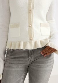 White and cream cardigan with a ruffled hem, featuring buttons and pocket details. Worn with gray jeans. Soft, textured fabric.