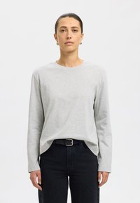 Grijs shirt met lange mouwen van zacht materiaal en een ronde hals. Draagt donkere spijkerbroek met een zwarte riem met zilveren gesp.