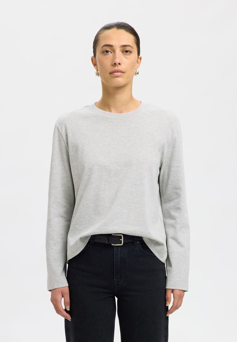 Grijs shirt met lange mouwen van zacht materiaal en een ronde hals. Draagt donkere spijkerbroek met een zwarte riem met zilveren gesp.