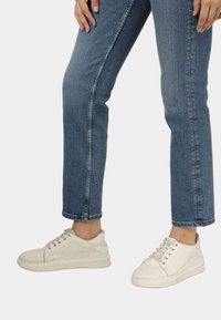 Ténis brancos com sola plana, textura lisa e perfurações subtis, combinados com jeans azuis com bainha.