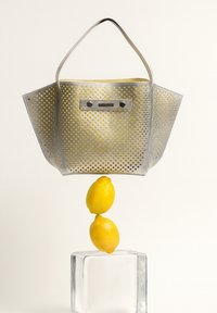 Metalen zilveren tote bag met gele voering, met een geperforeerd ontwerp en twee handvaten, opgehangen boven twee opgestapelde citroenen op een heldere kubus.